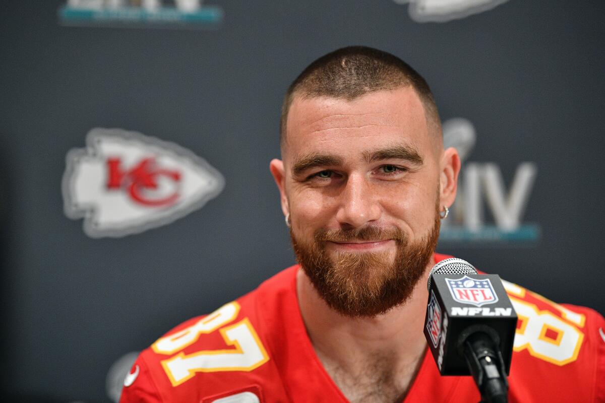 Travis Kelce sits at a desk with buzzed hair and small silver earrings in an orange jersey