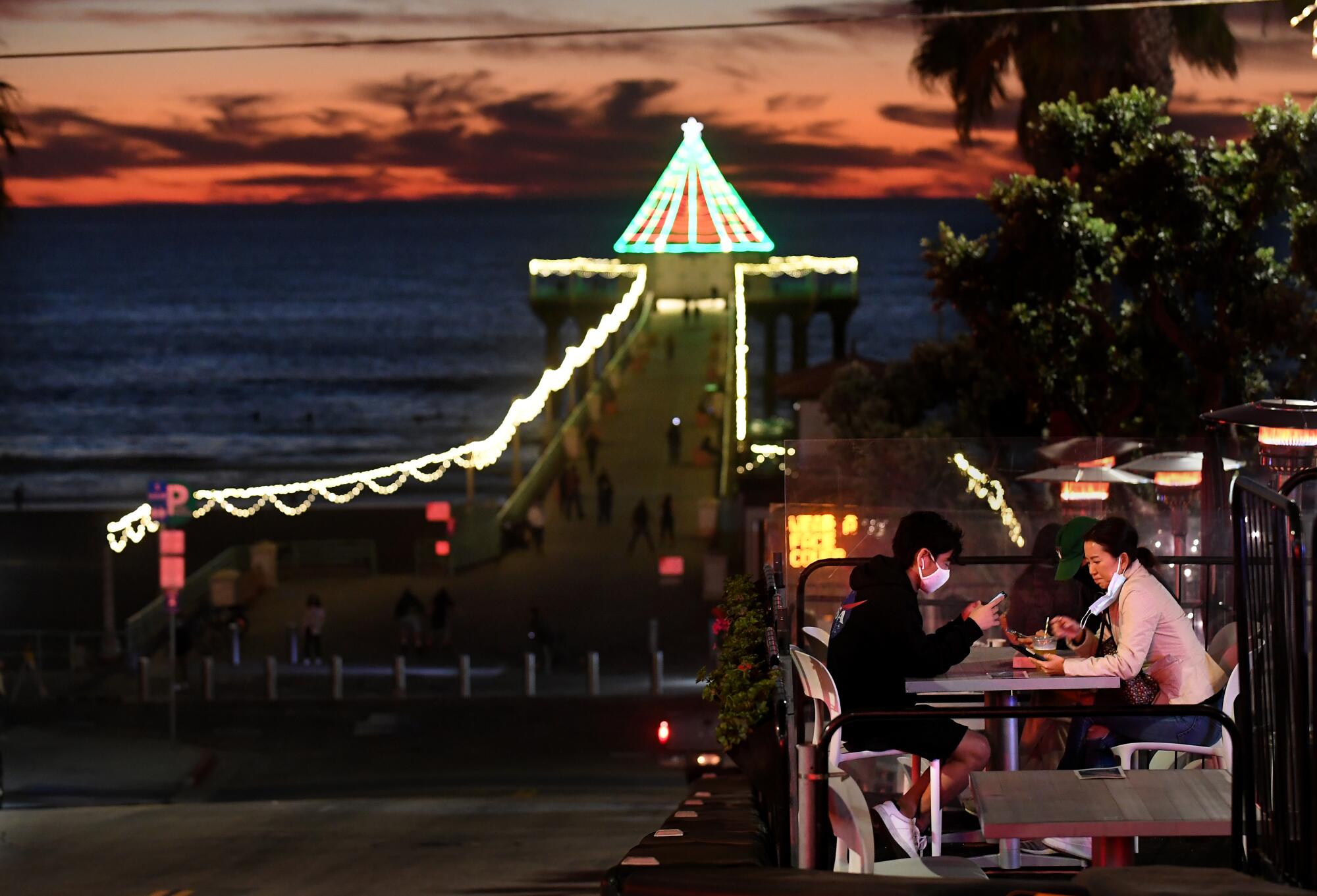 People dine in the early evening next to a pier decorated with holiday lights.