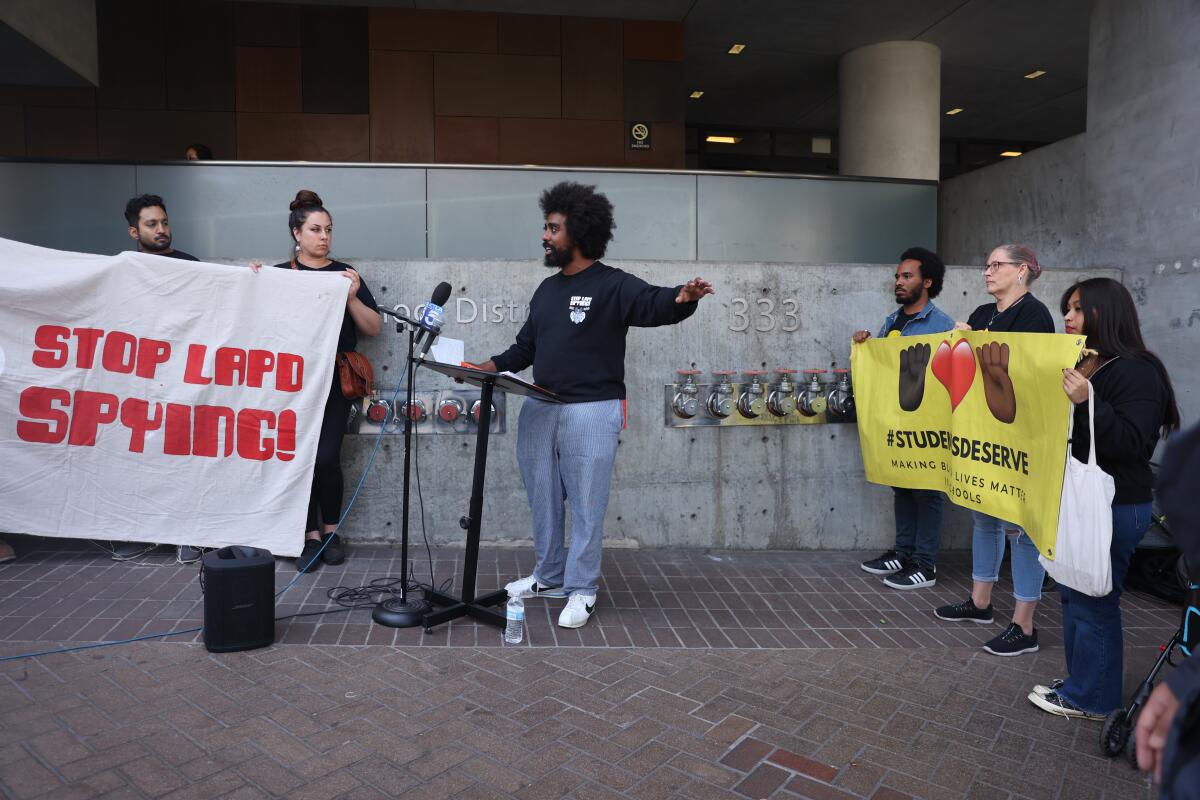 A man with dark hair, in a dark shirt, speaks while he is flanked by people holding banners. One says Stop LAPD Spying 