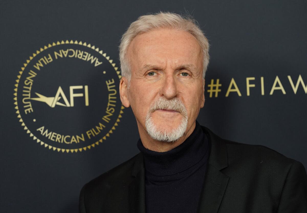 James Cameron poses in a black turtleneck and black suit against a black-and-gold background.