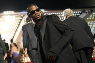 Travis Scott looks over his shoulder in black sunglasses, suit and gloves