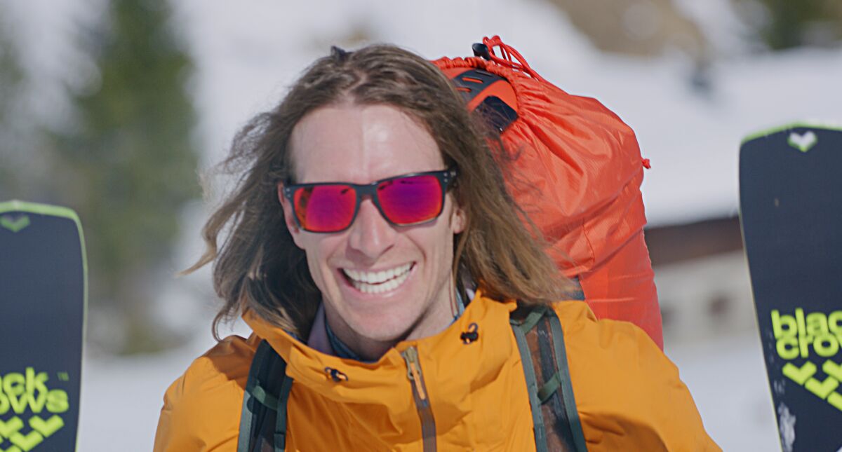 A long-haired outdoor athlete carries gear on his back.