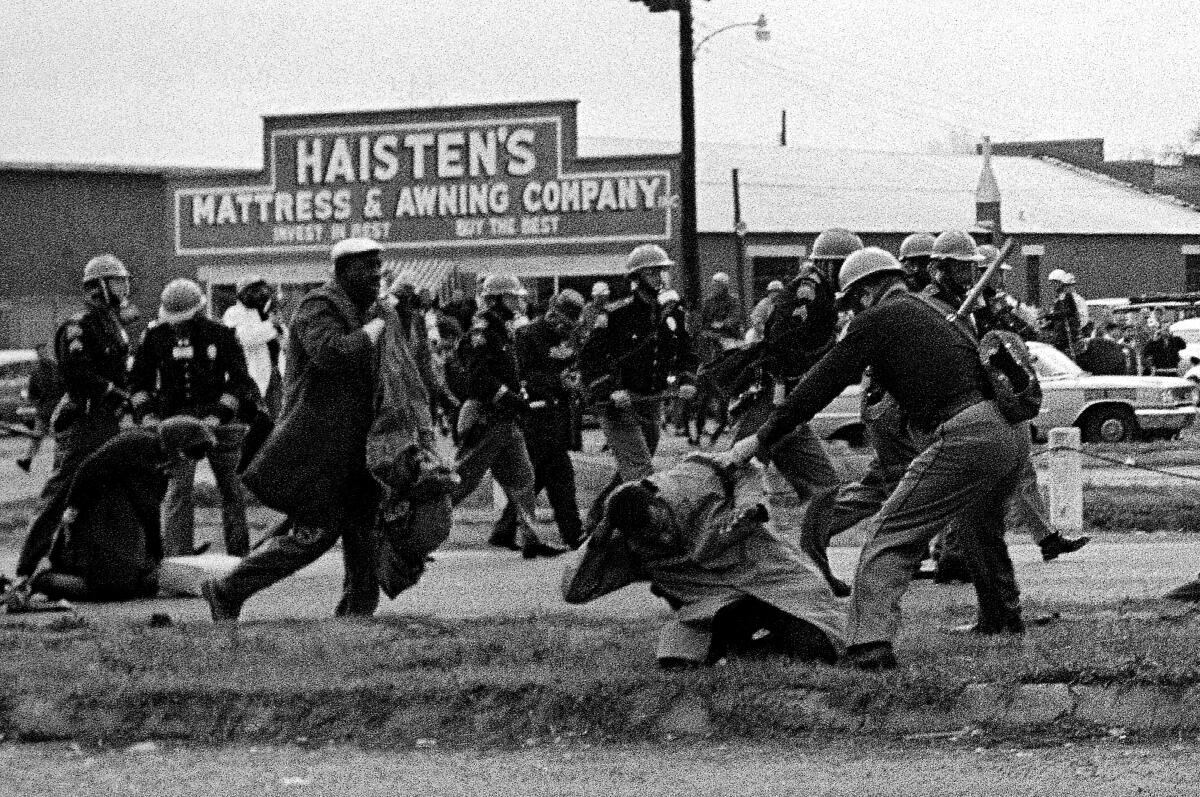 A man kneels, trying to cover his head, as a man in uniform swings a club at him. Other uniformed men swarm behind.