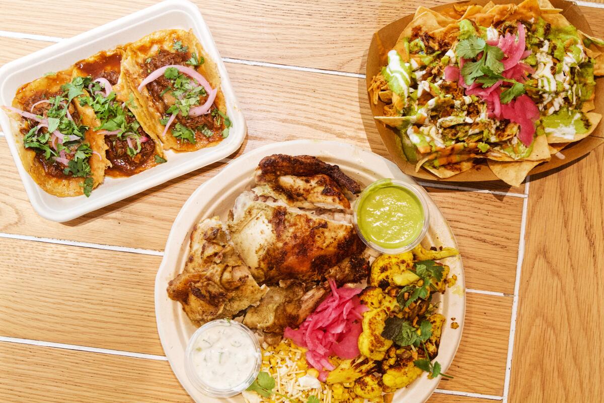 An overhead photo of three birria tacos, a rotisserie chicken plate and pibil chicken nachos atop a wood tabletop.