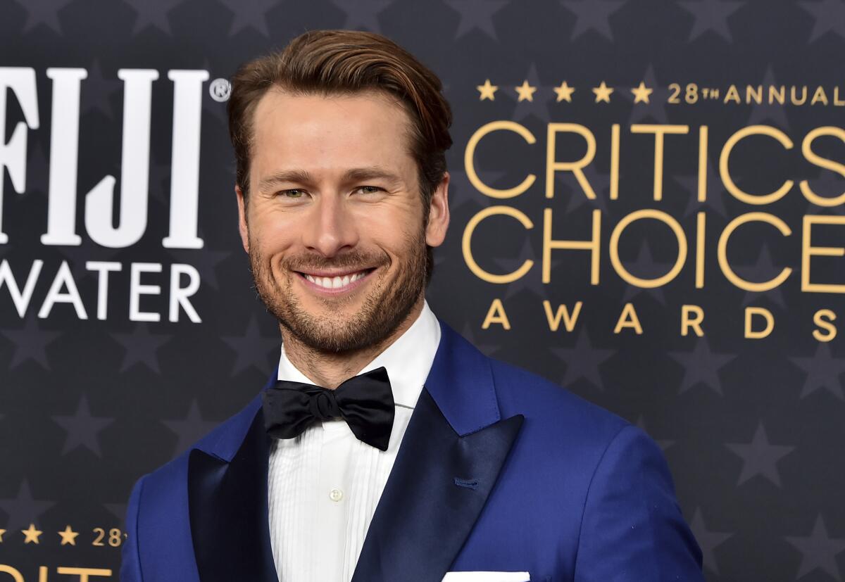 Glen Powell wears a blue tuxedo with a black bowtie and a white tux shirt as he smiles broadly