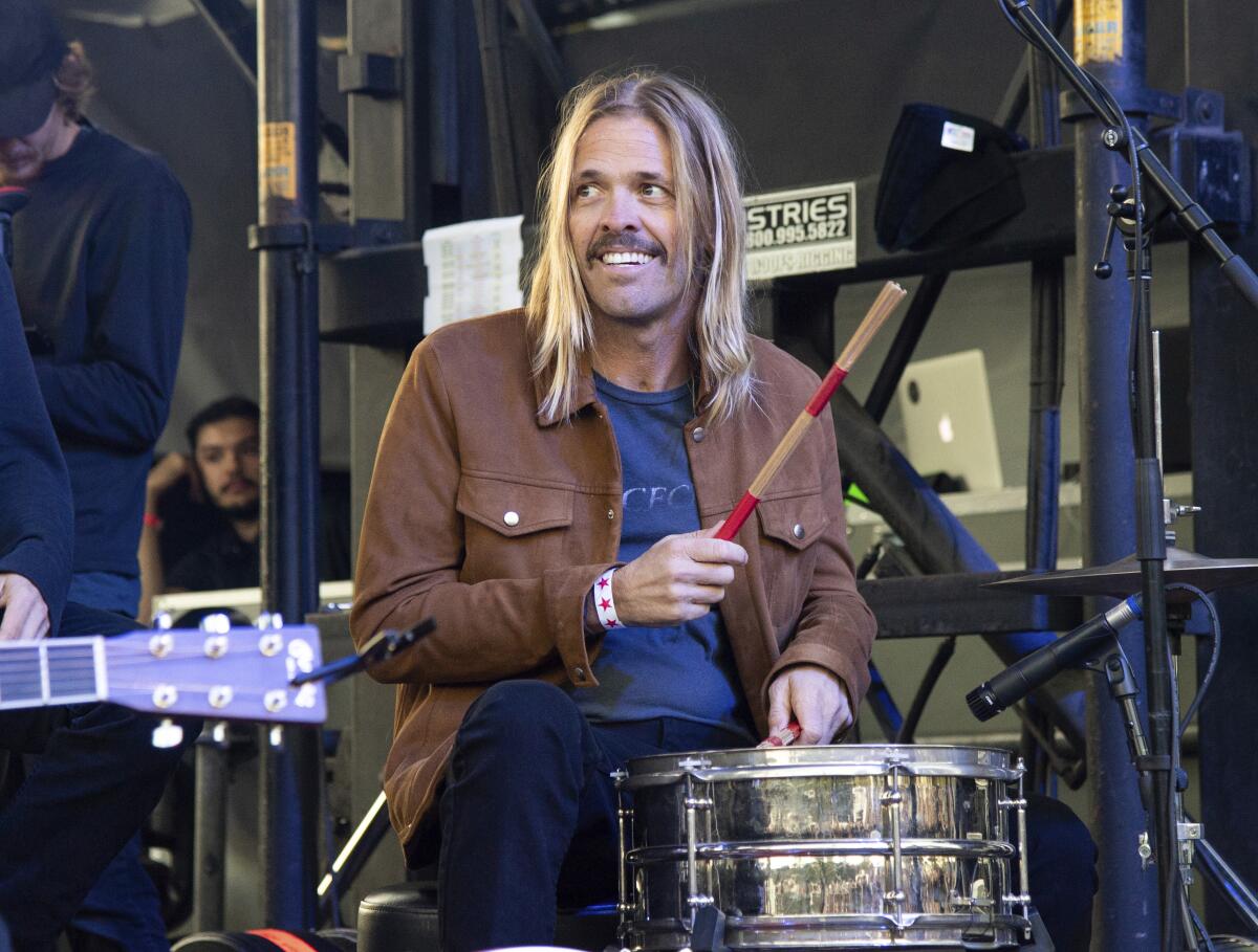 A man with blond hair wearing a brown jacket plays the drums on stage