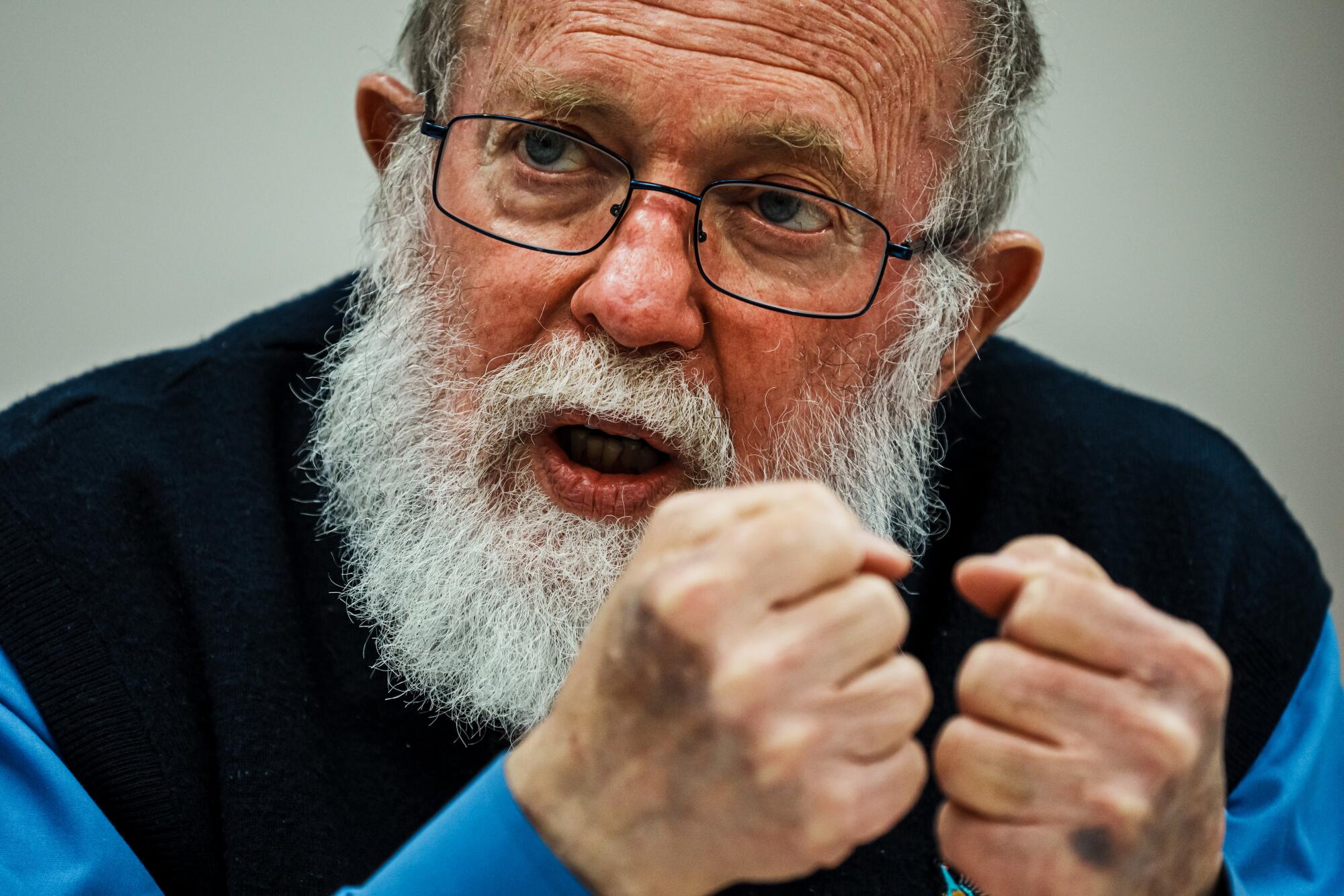 A closeup of a man with glasses and a white beard, holding his fists together near his face as he speaks