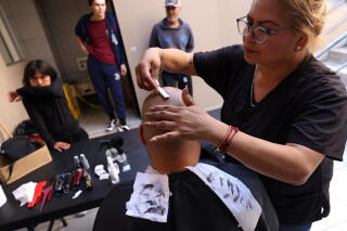 A woman in dark shirt runs a blade over the head of a bald man seated with strips of paper over his back, with hair on them