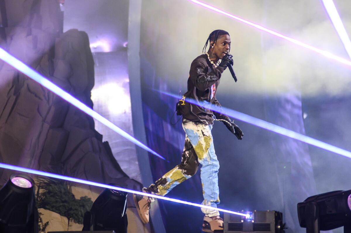 Travis Scott walking across a concert stage rapping into a mic and wearing trashy jeans, a black T-shirt and black gloves. 