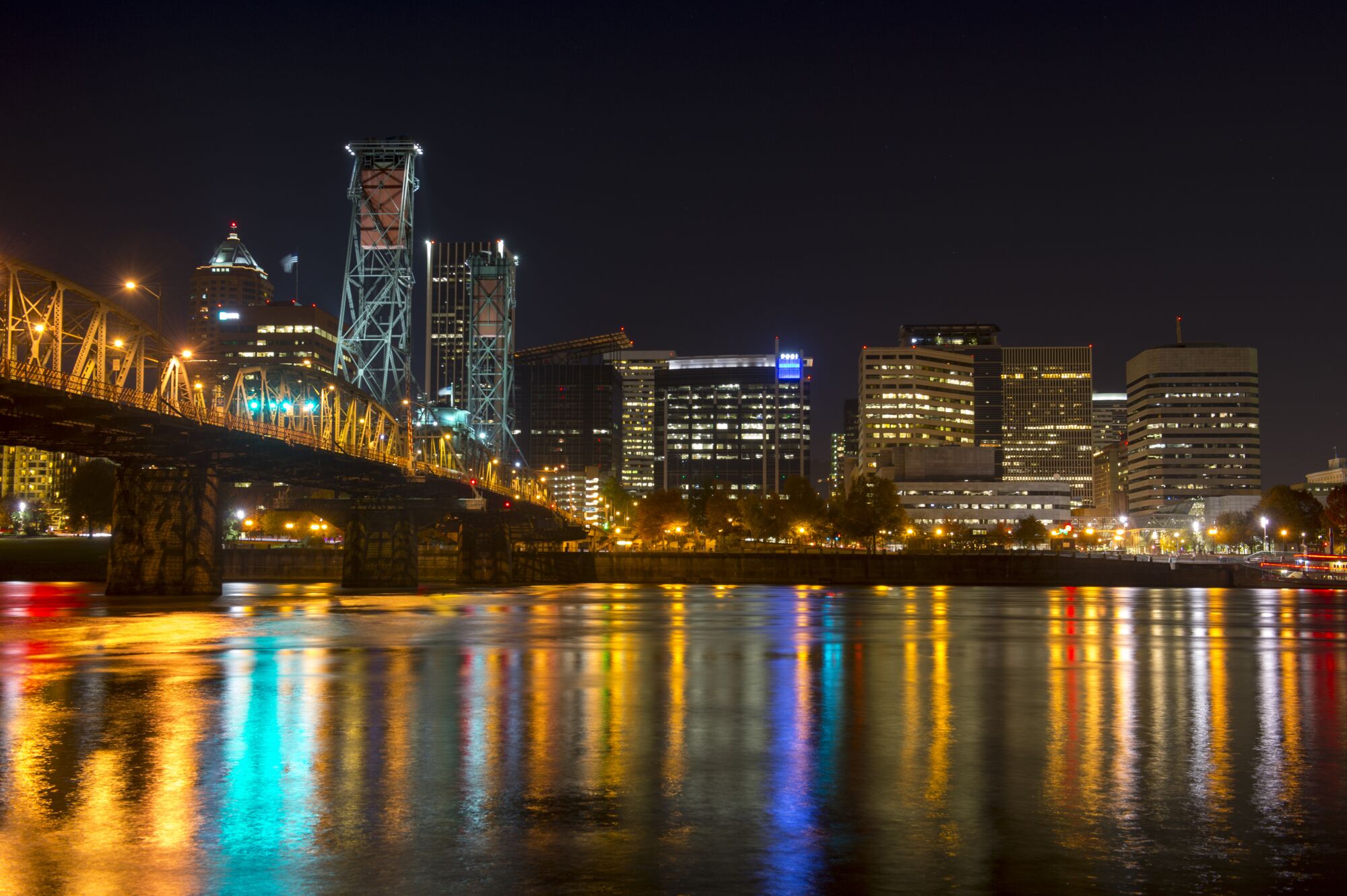 The lights of the downtown Portland skyline, seen at night next to the Hawthorne Bridge, reflected by the Willamette River