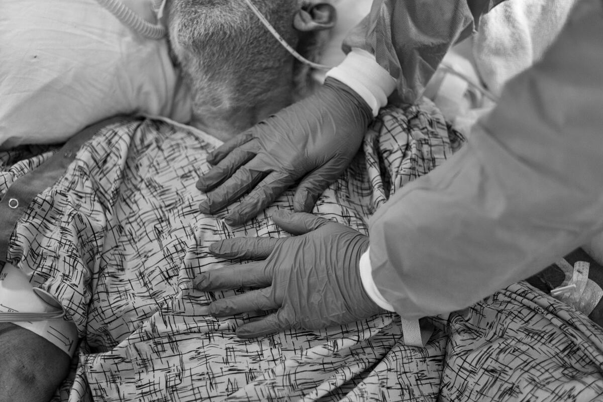 The gloved hands of a chaplain hover over the heart of a COVID-19 patient with oxygen tubes in his nose.