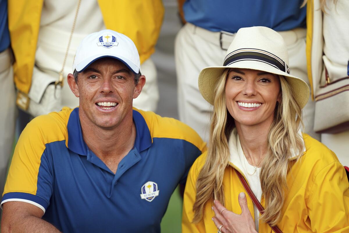 Rory McIlroy in a cap and blue golf shirt with yellow trim sitting next to wife Erica Stoll in a yellow windbreaker and hat