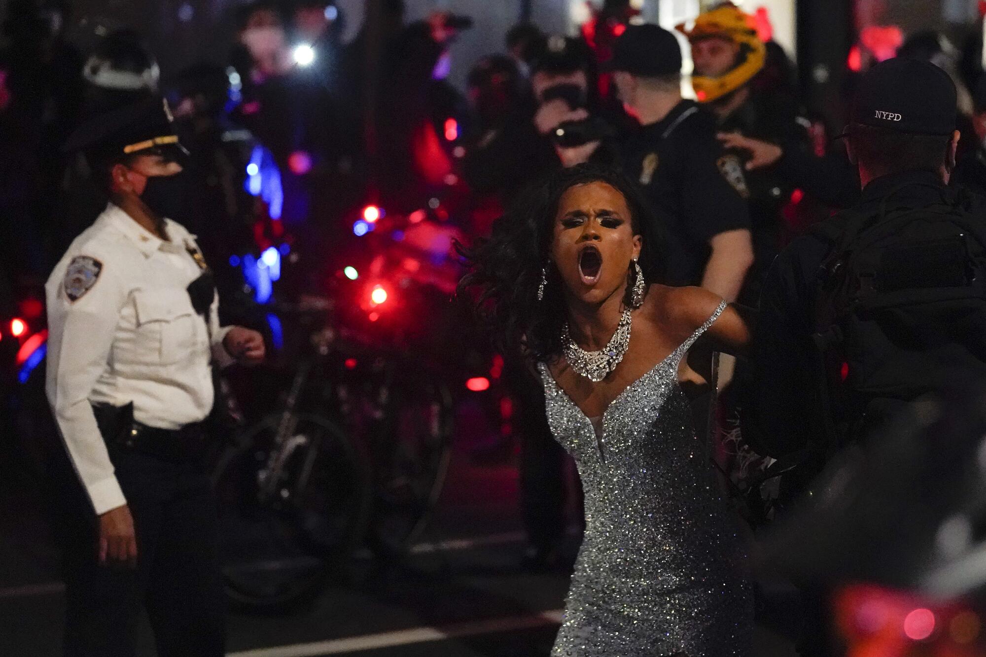 A drag queen in a silver sequined dress yells as they are arrested by police