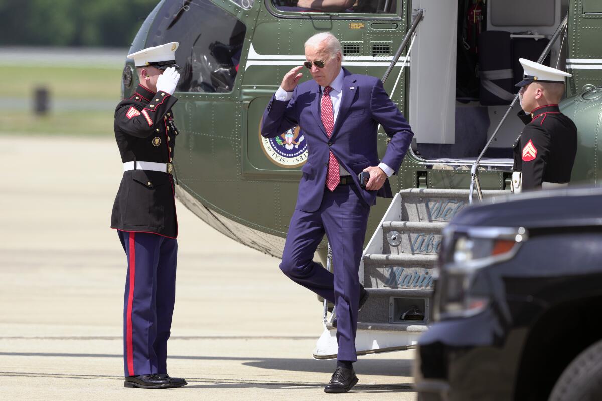 President Biden walks down the steps of a helicopter and salutes two service members. 