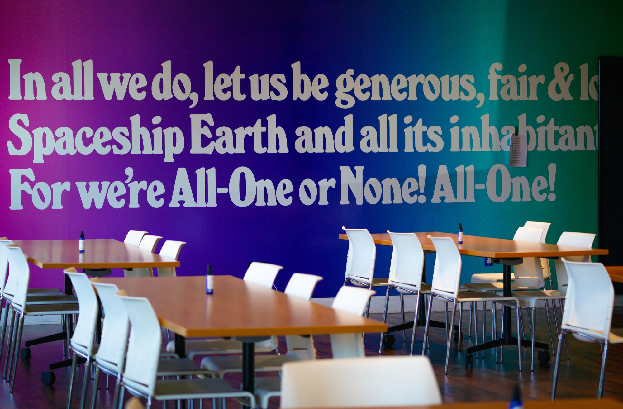 Empty tables and chairs in front of a colorful mural with a saying that reads in part, "In all we do, let us be generous."