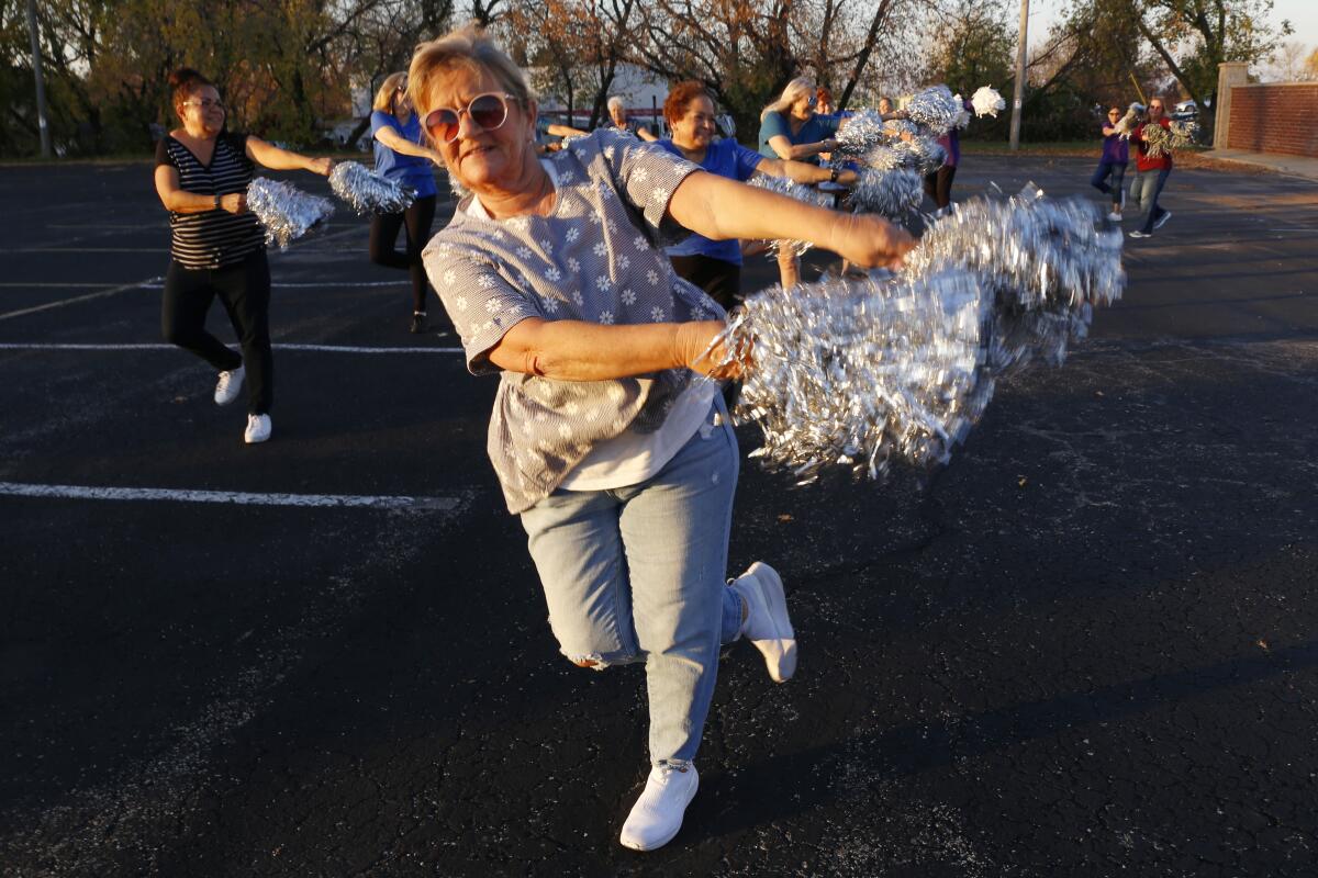 Older women dance in a parking lot with silver pom poms 