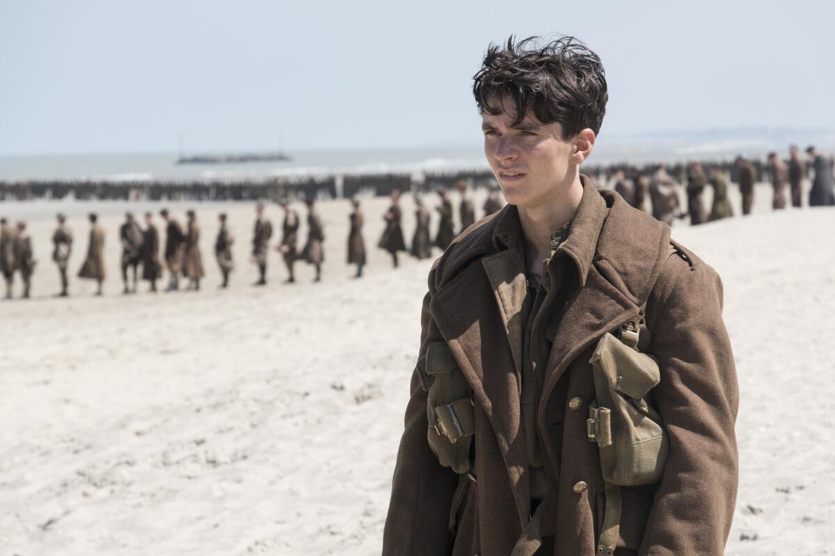 A young soldier stands alone on the beach, a line of men behind him.