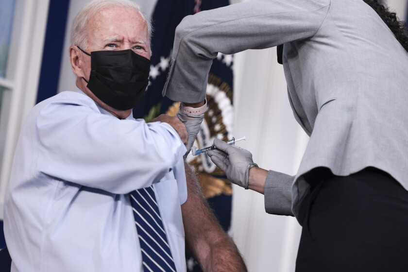 An older man in a mask, dress shirt and tie holds up his sleeve while receiving an inoculation.