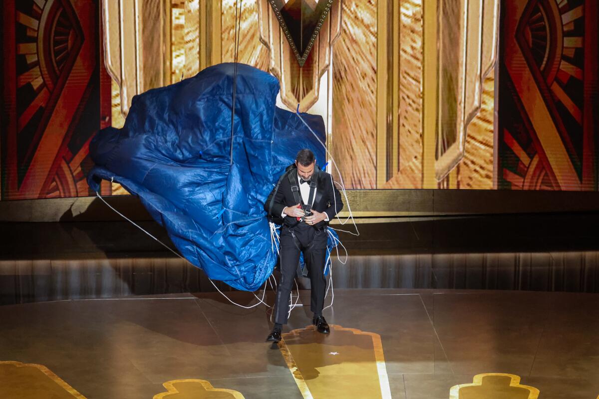 Jimmy Kimmel, in a black tuxedo, is looking down and is unbuckling a large blue parachute that is floating behind him.