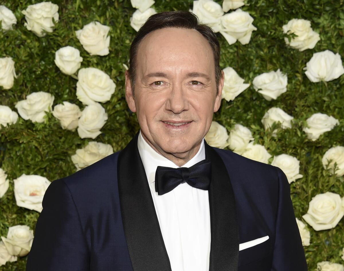 Kevin Spacey smiles in a tuxedo in front of roses and greenery.