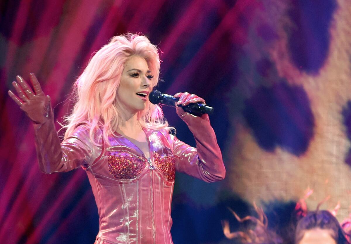 A blond woman onstage, dressed in a pink leather cowboy jumpsuit, holds a microphone to her mouth