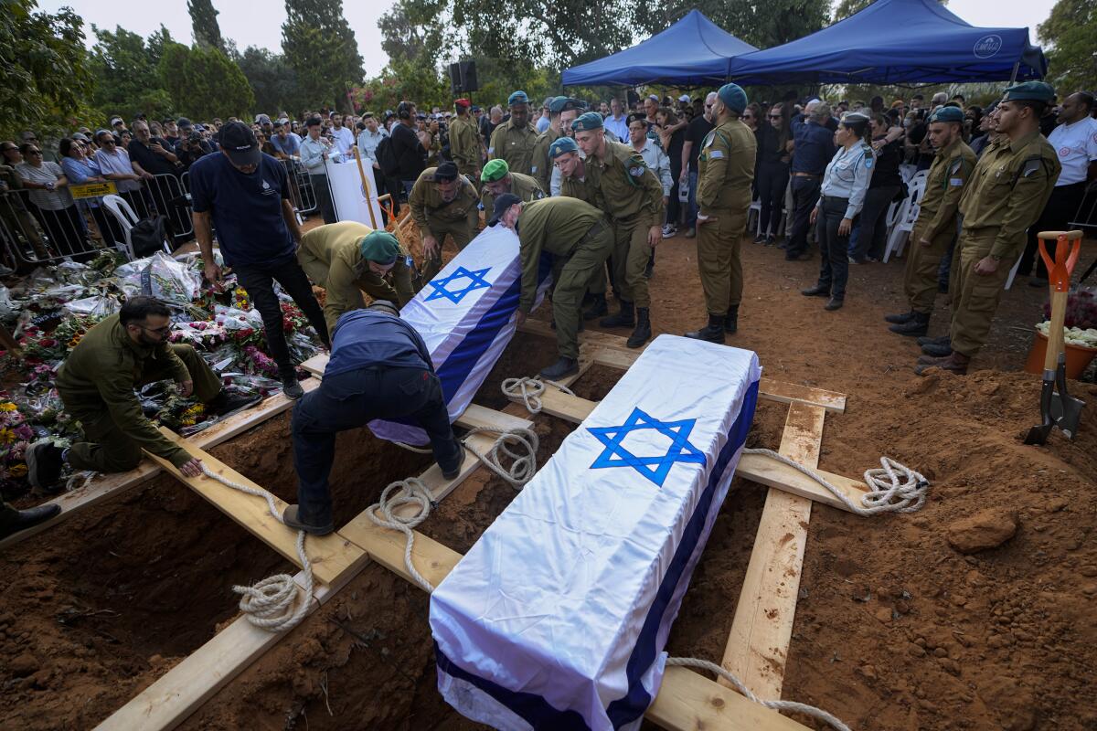Soldiers put a coffin covered in Israel's flag next to another on a wood platform over two fresh graves as a crowd looks on.