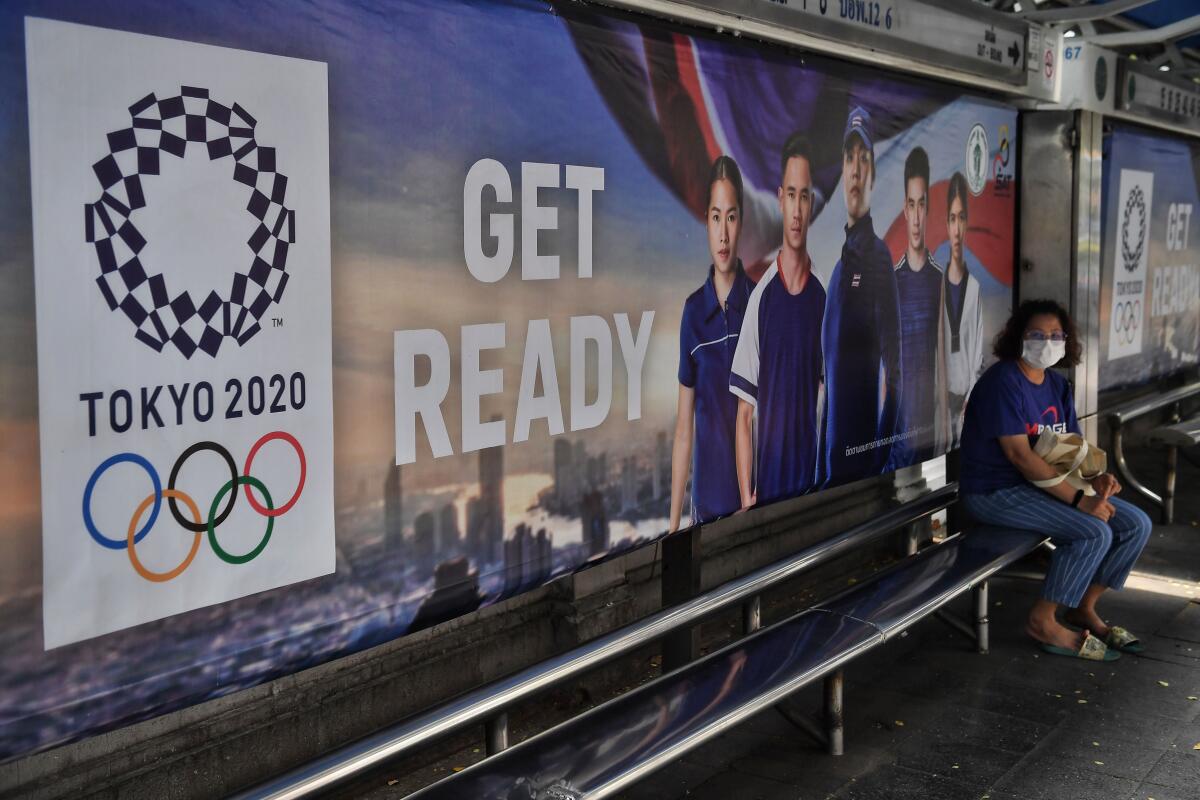 A woman in a mask in Bangkok sits in front of a sign for the 2020 Olympics.