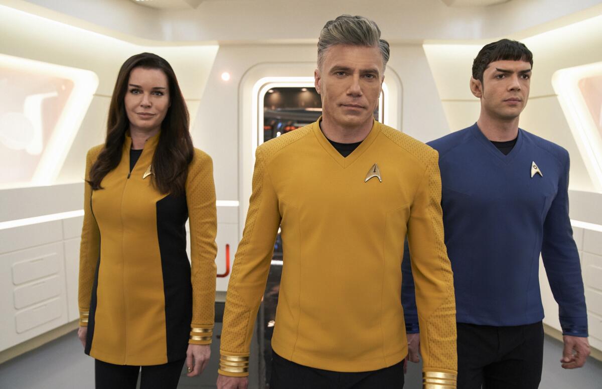 A woman and two men in yellow and blue uniforms walk through a white hall in a spaceship.