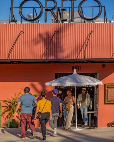 People walk to an orange building, its entrance under an umbrella and the word Loreto in tall black letters atop the building