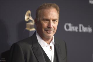 Kevin Costner posing at an angle in a black blazer and white suit shirt