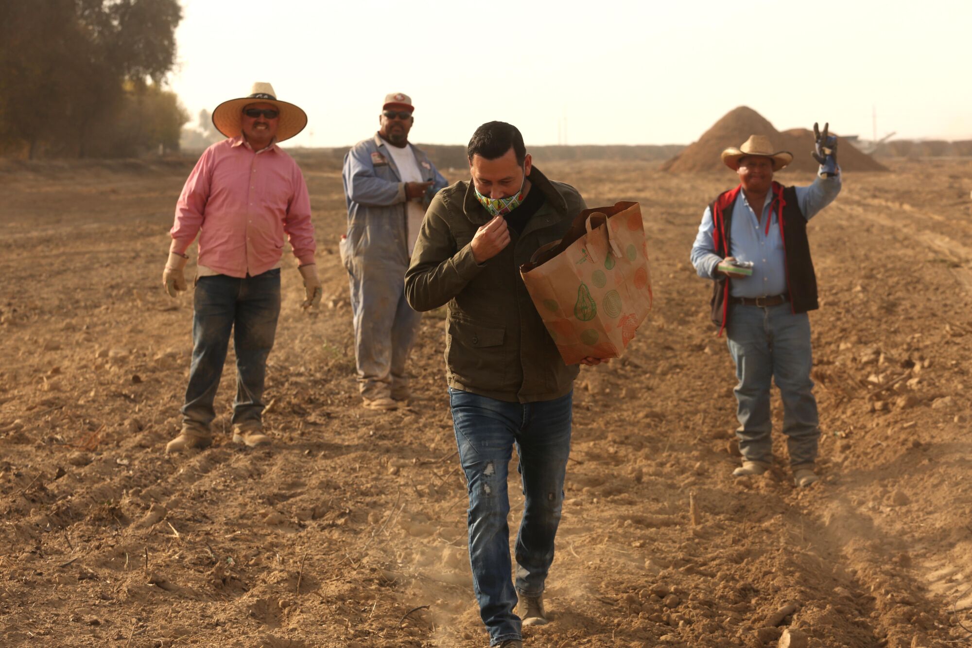 Men in a dusty, barren field wave to a man walking away from them holding a paper bag