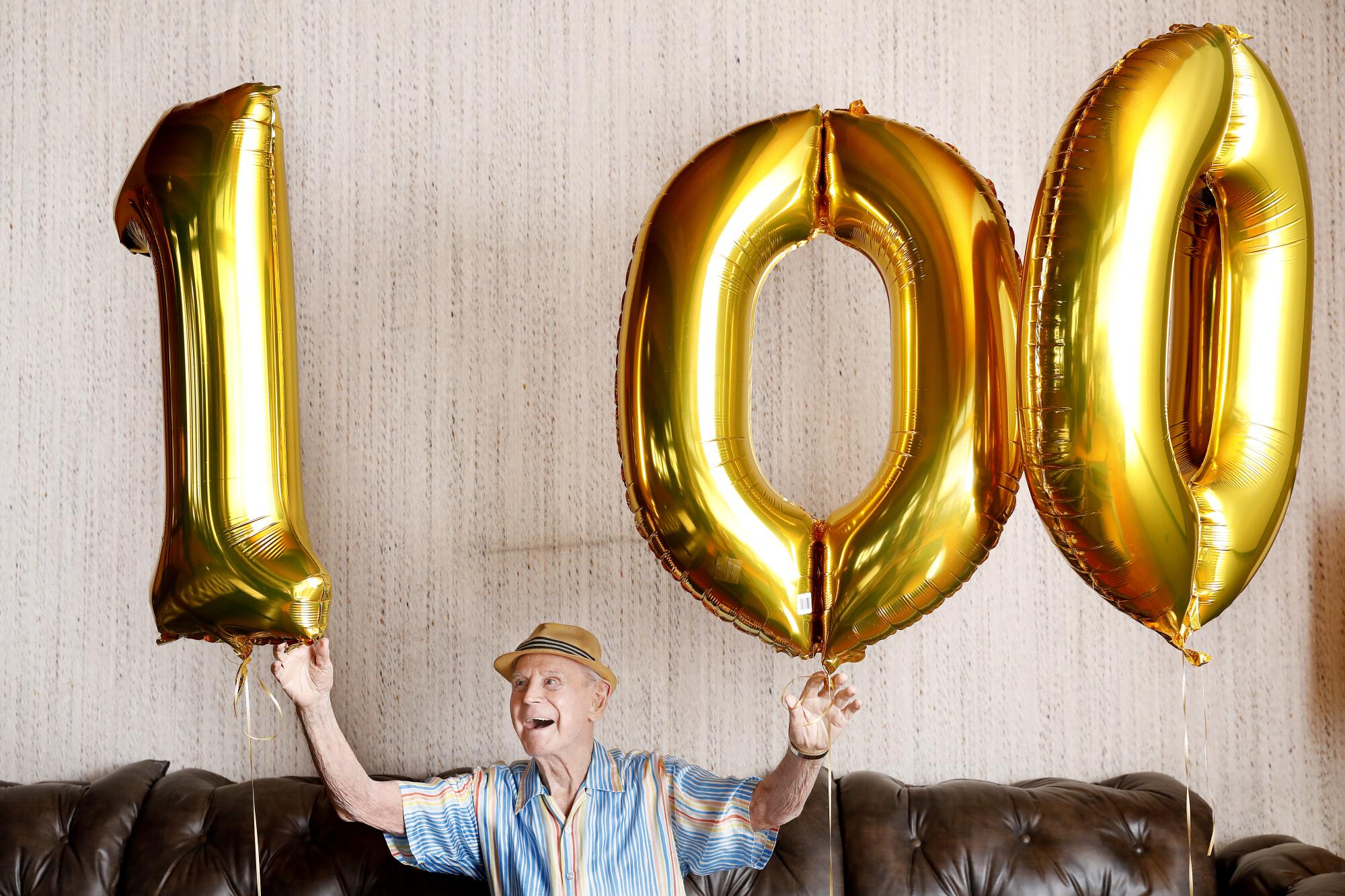 A man in a fedora holds up 3 balloons that spell "100" 