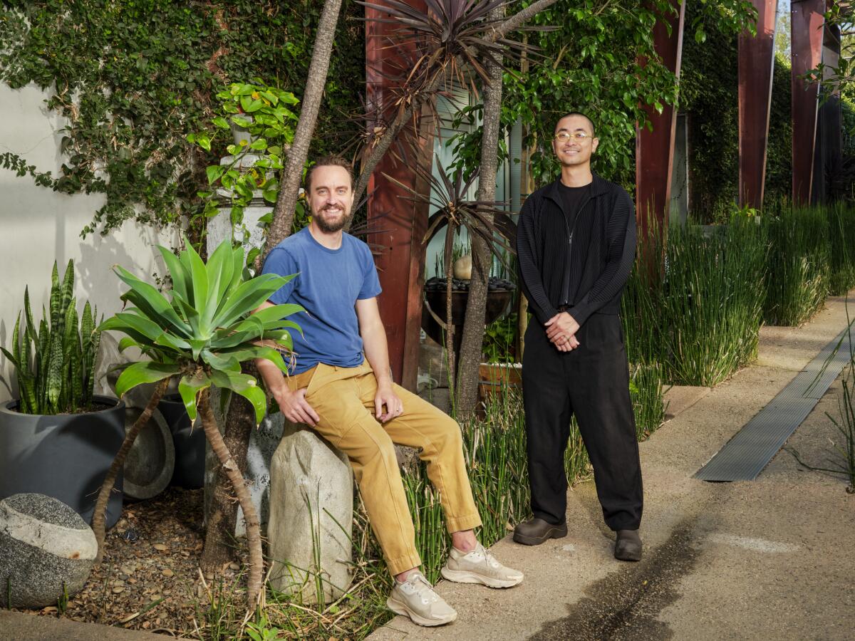 One man sits on a rock amid plants, and another stands. Both are smiling