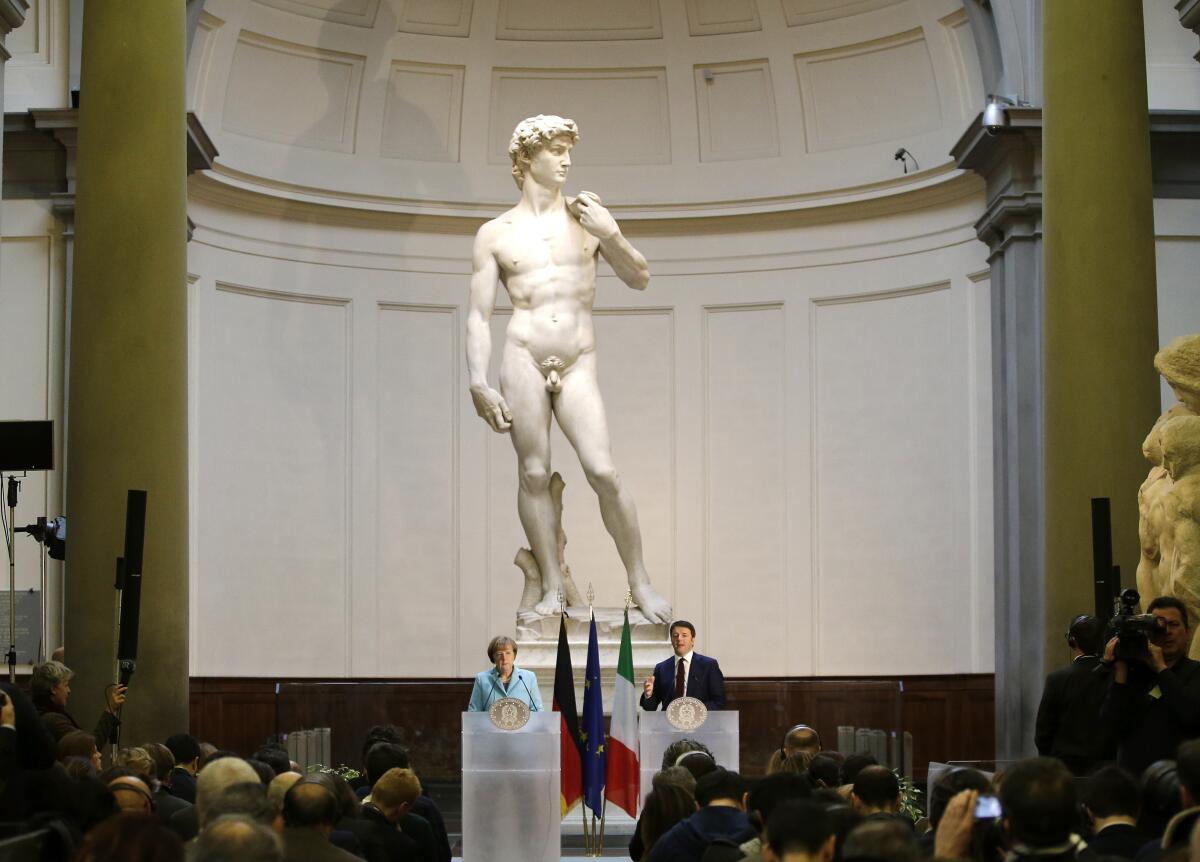 A man and woman dwarfed by Michelangelo's "David" as they speak to a seated audience from in front of the statue.