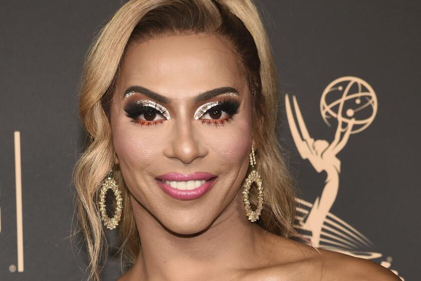 A person with light blonde and brown hair wearing a gold one-shoulder dress smiles