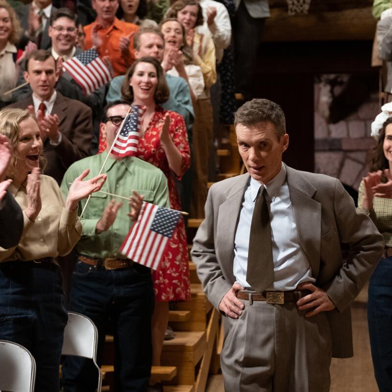 A man in a 1940s suit walks among people clapping and waving small American flags