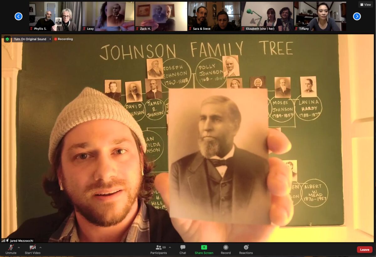A man holds a black and white photo of his ancestor in front of a chalkboard depicting a family tree