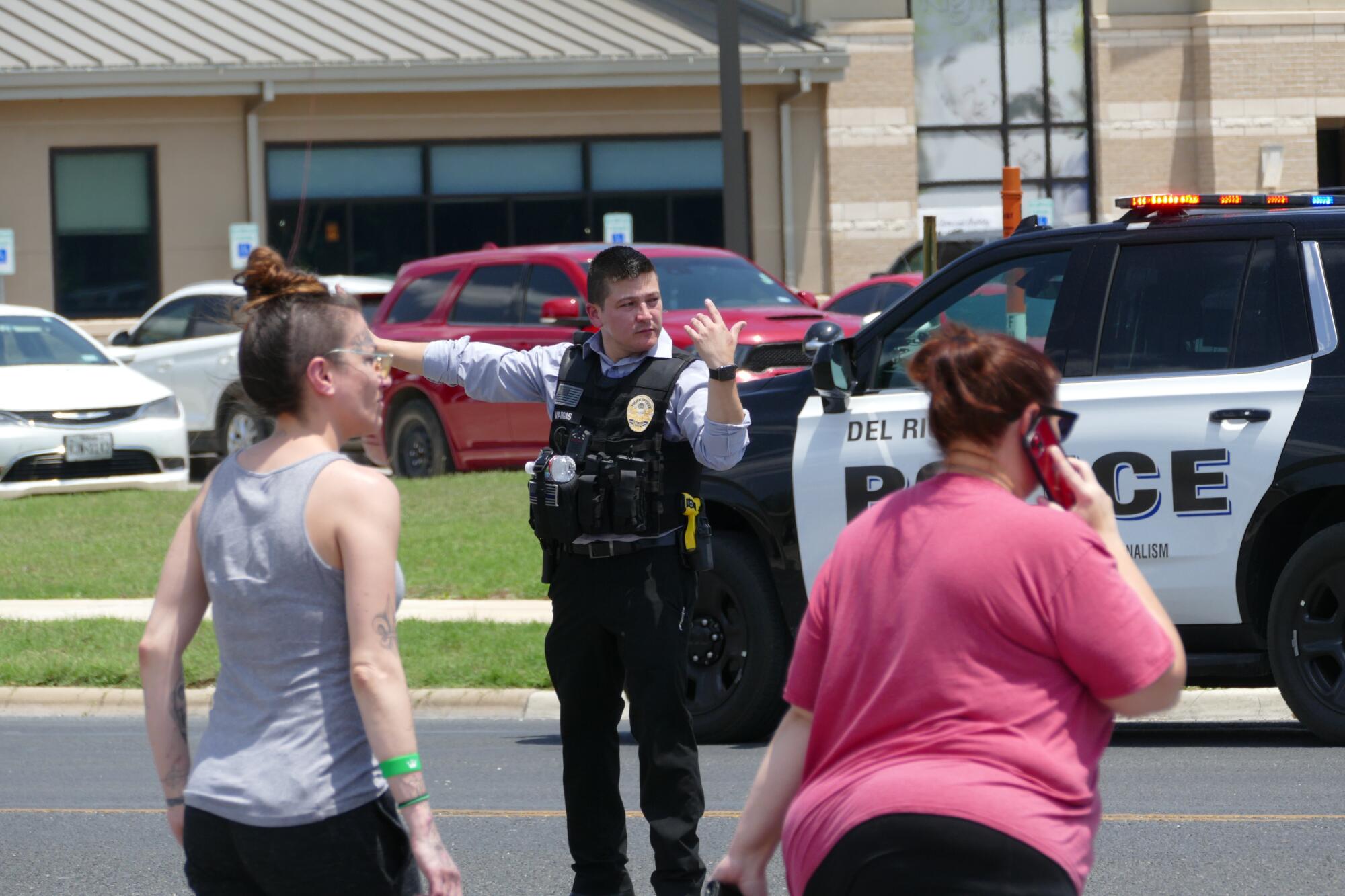 An officer in uniform gestures as he stands in front of a police vehicle facing two women crossing a street 