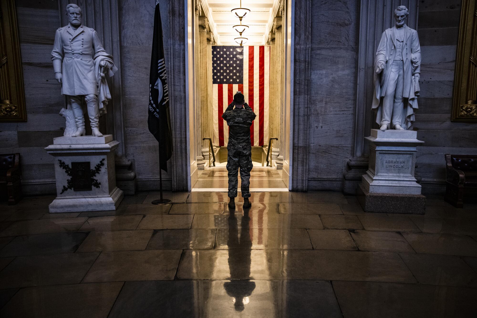 A troop in camo fatigues stands in front of a large flag in the Capitol 