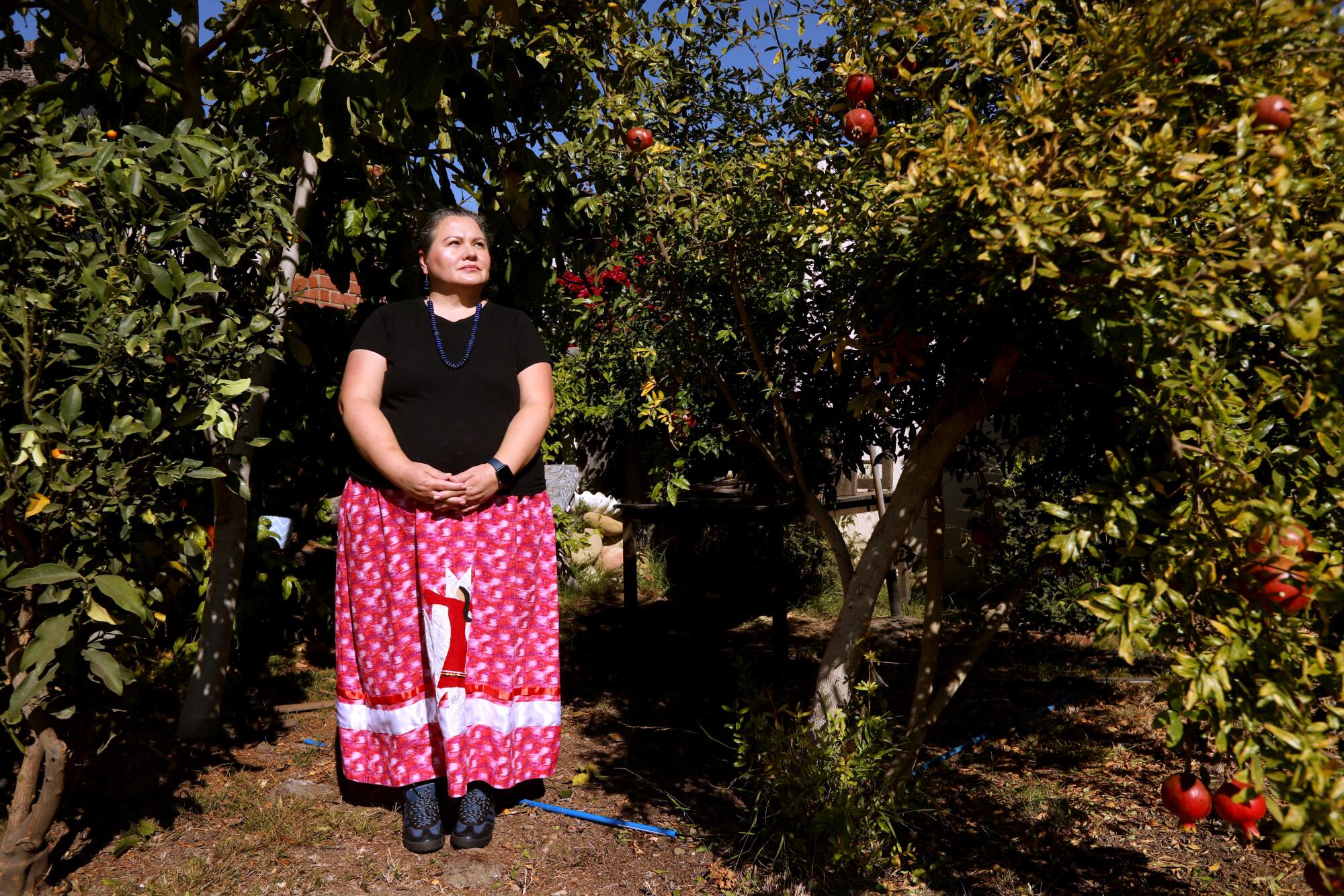 A woman in a black top and red-and-white patterned skirt stands near a pomegranate tree with hands clasped