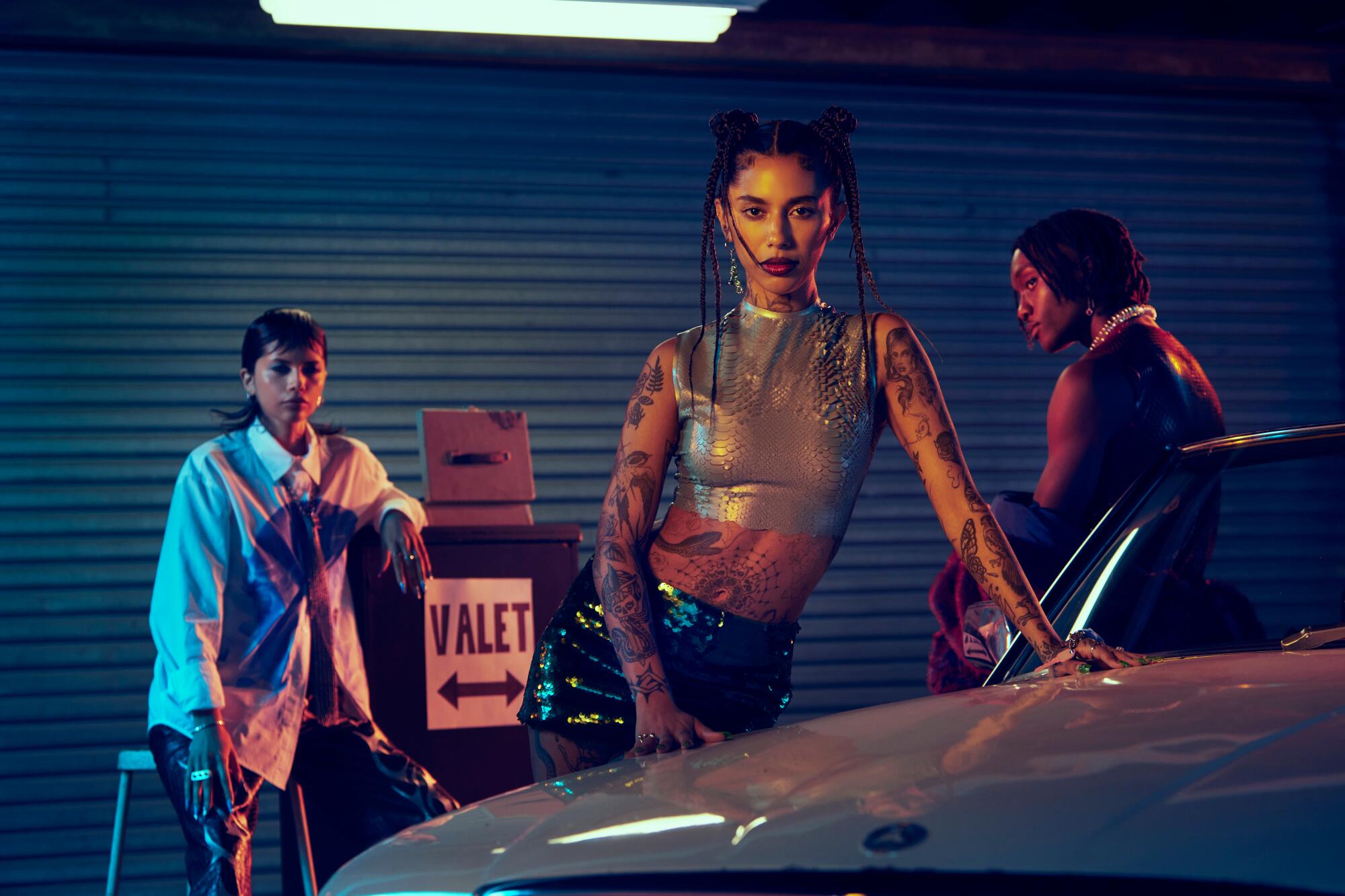 A tattooed woman leans against the hood of a car, while two figures in the background stand beside a "valet" sign.