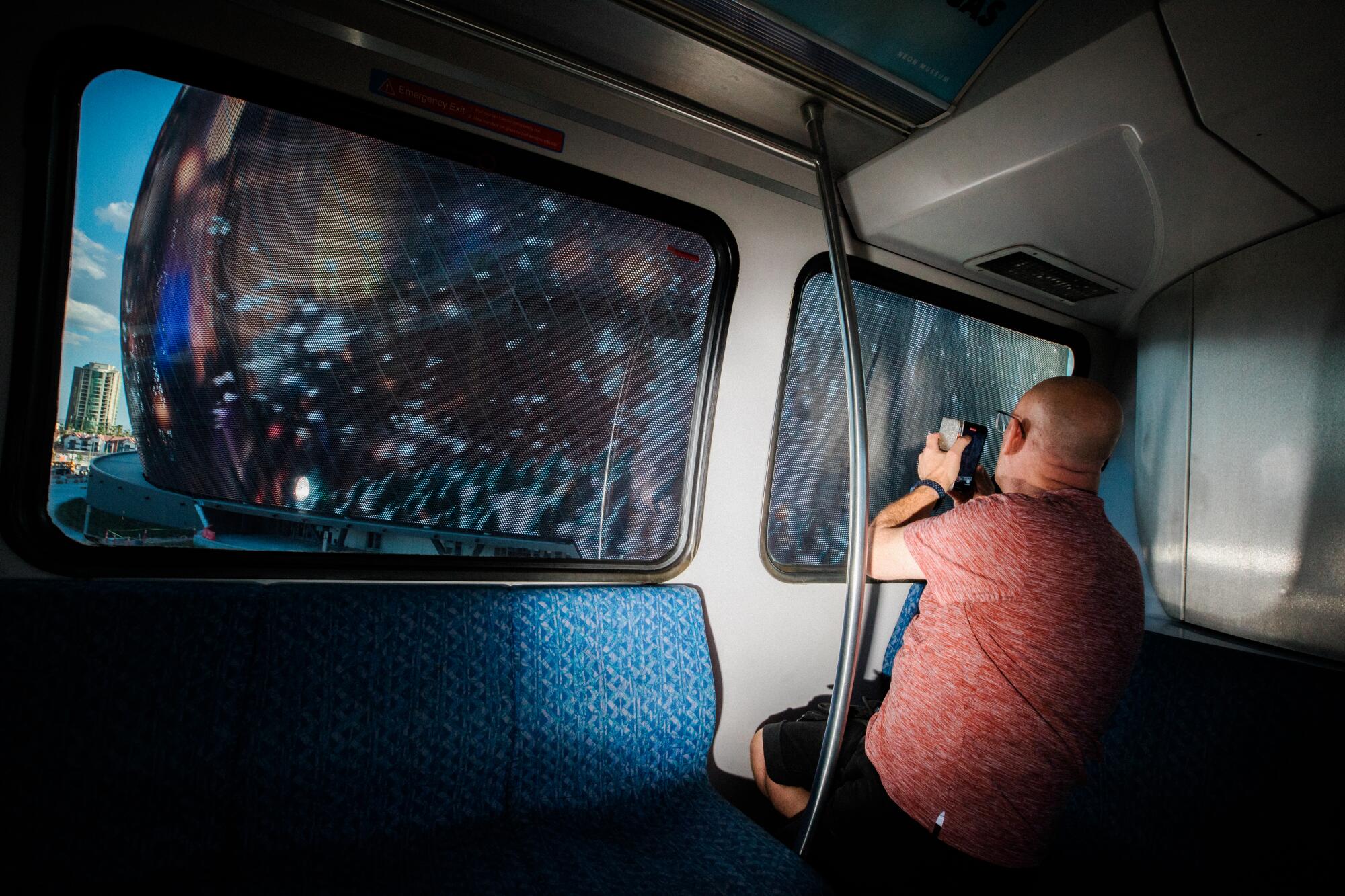 A man in a red shirt holds up a smartphone to record Sphere from a monorail car. 