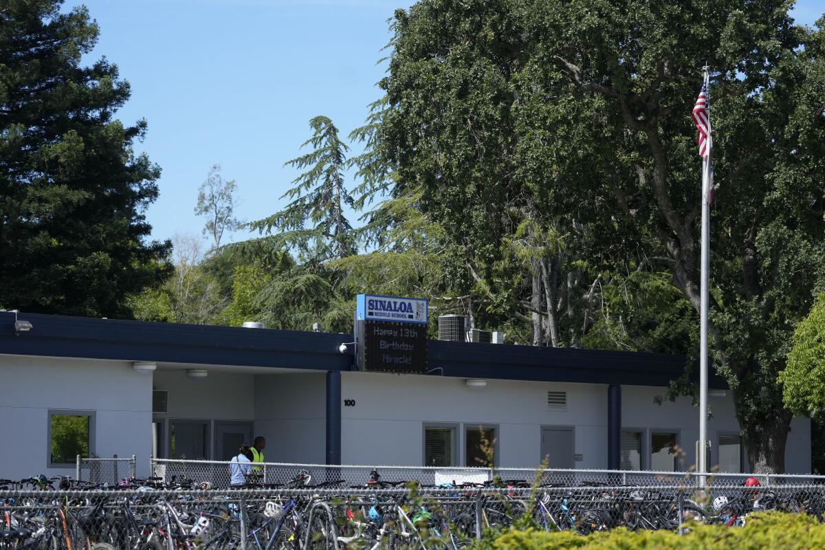 Bicycles are parked in front of a long, low school building surrounded by trees.