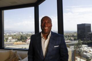 Byron Allen smiling in a dark blue suit in an office with a window view overlooking Los Angeles