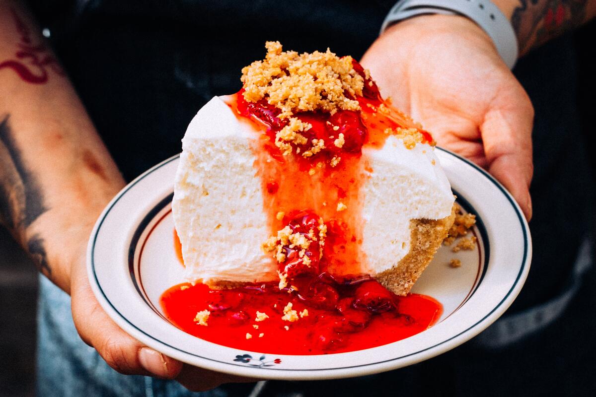 Hands hold out a plate with a sliced of whipped cheesecake with a red sauce.