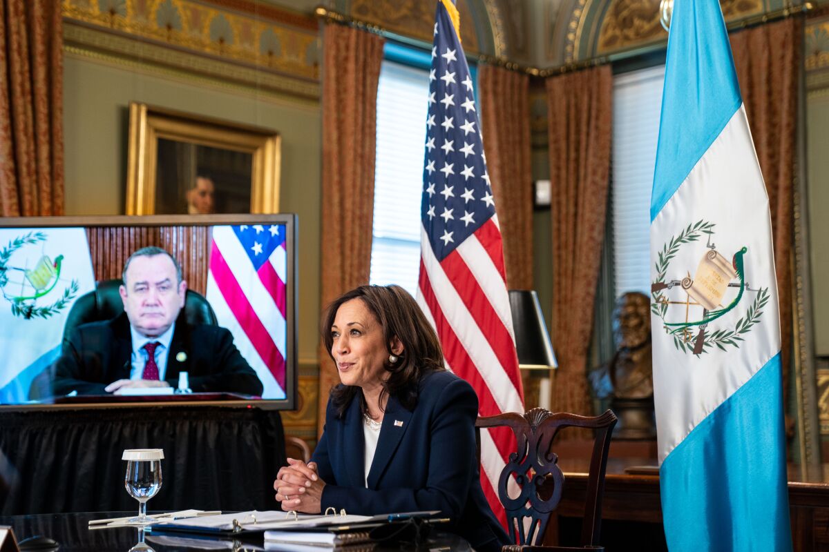 Kamala Harris sits attentively at a desk while a man in a suit appears on a screen next to her. 