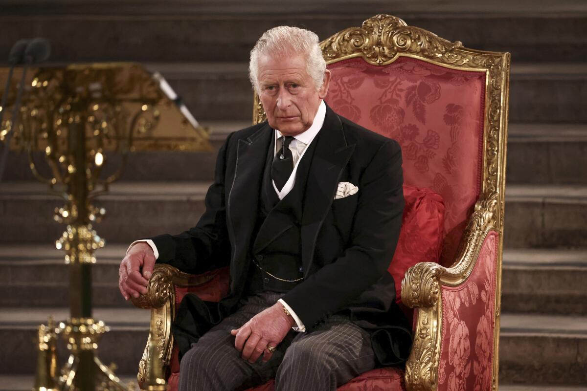 A gray-haired, solemn-looking man in a dark suit and tie sits in a gilded chair with red damask upholstery 