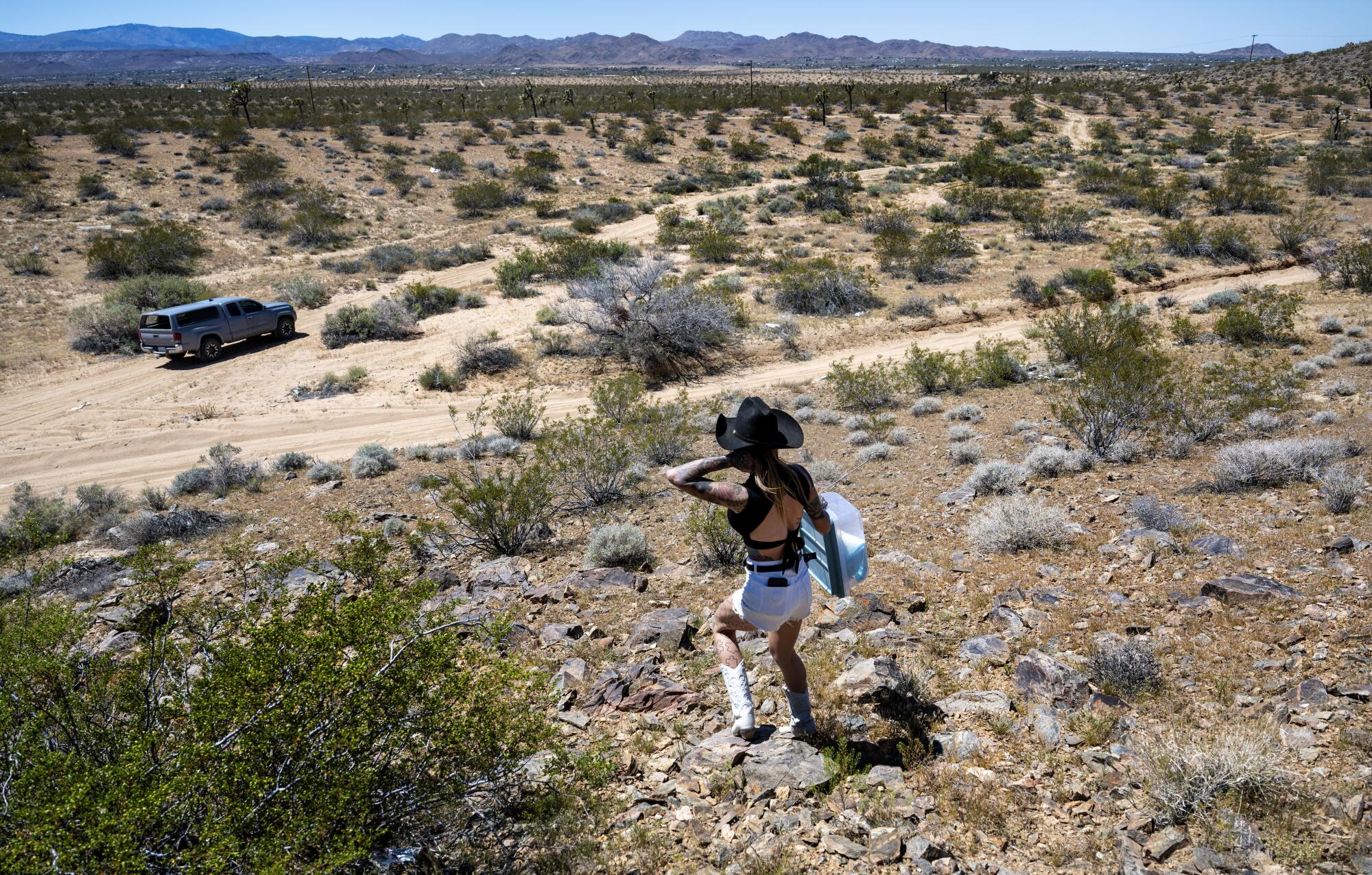 Wall trudges down a hill toward her truck in the high desert after relocating a snake. Jagged hills rise in the distance.
