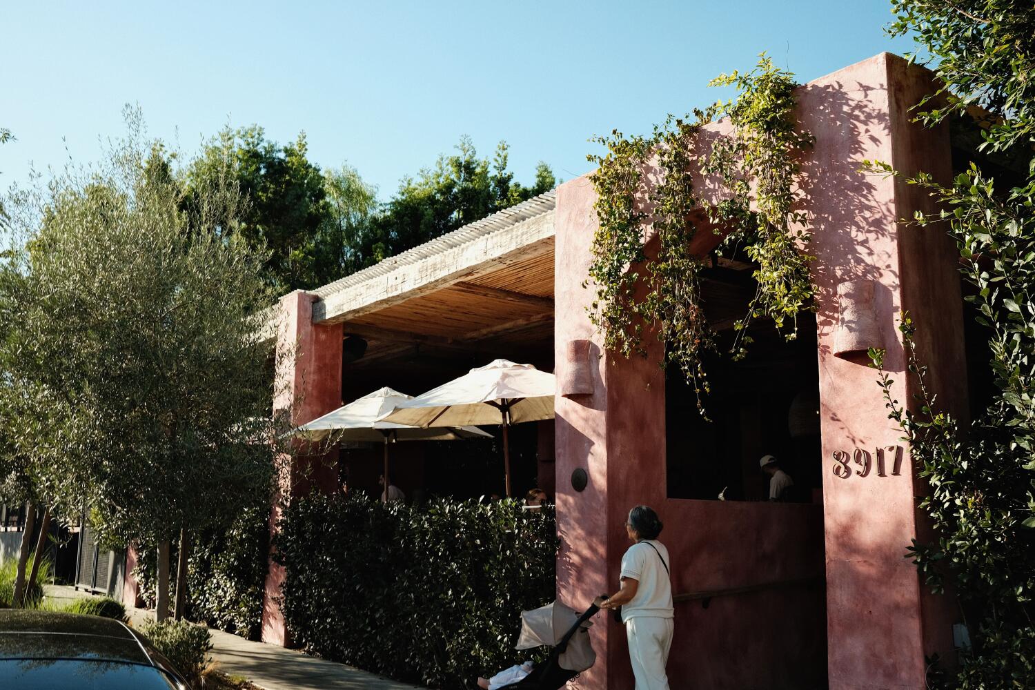 An exterior of a light-pink building dotted with foliage. Near the entrance, a woman pushes a stroller.