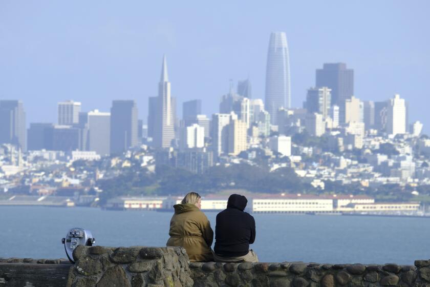 A couple sits at a vista point with the San Francisco skyline in the background Friday, March 27, 2020, in Sausalito, Calif. The surge of coronavirus cases in California that health officials have warned was coming has arrived and will worsen, Gov. Gavin Newsom said Friday, while the mayor of Los Angeles warned that by early next week his city could see the kind of crush that has crippled New York.(AP Photo/Eric Risberg)